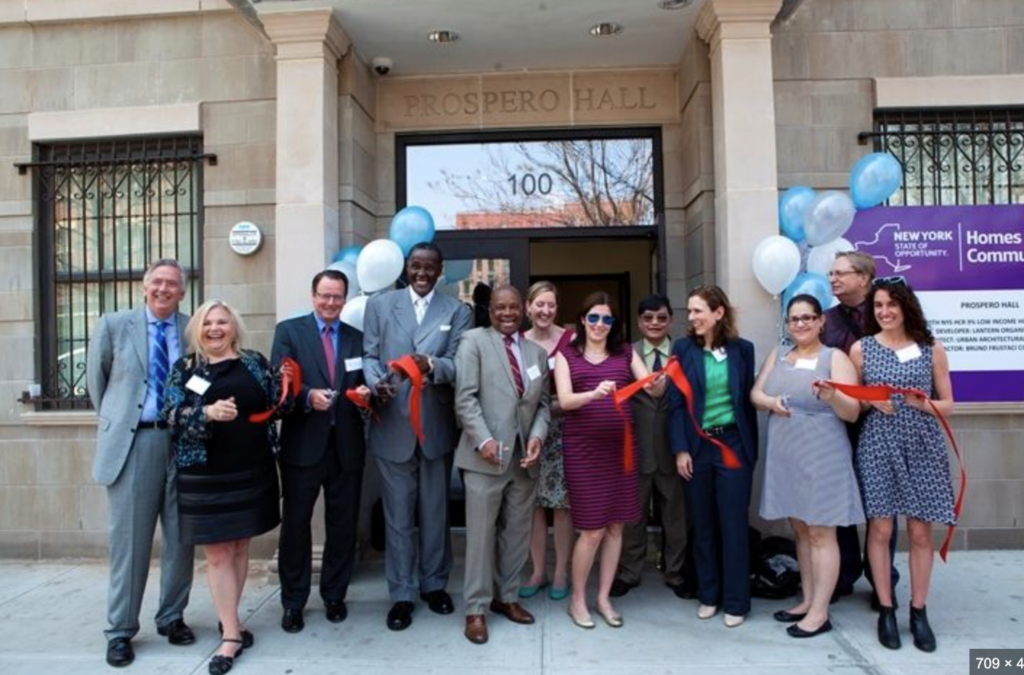 A group of eleven smiling individuals hold red ribbons and scissors at a ribbon-cutting ceremony for Prospero Hall.